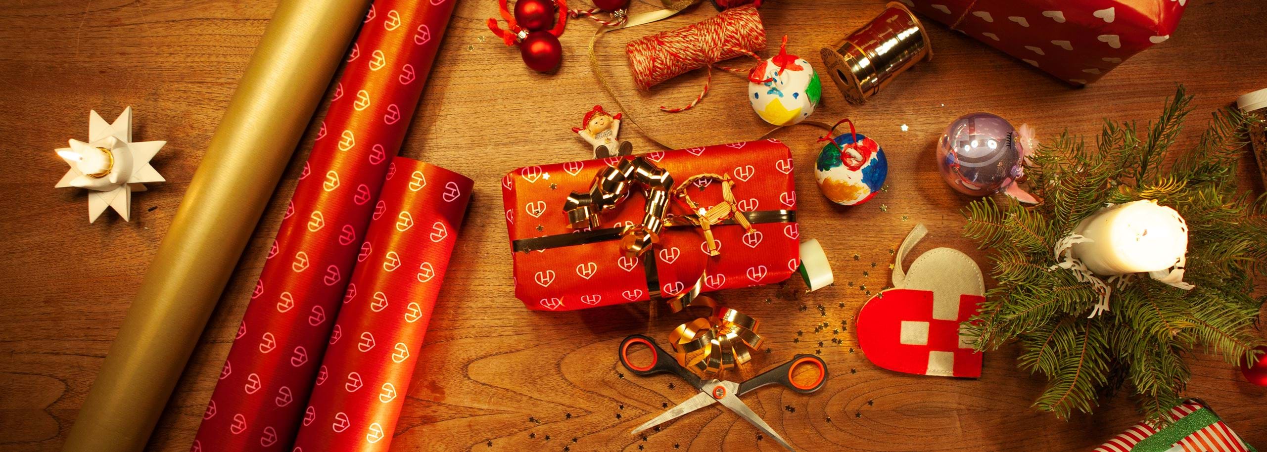 Overhead view of Christmas gift wrapping on a wooden table with red wrapping paper printed with the Hindenburg logo, a wrapped present, scissors, ribbon, festive ornaments, and a silver unicorn Christmas bauble featuring the Hindenburg logo.
