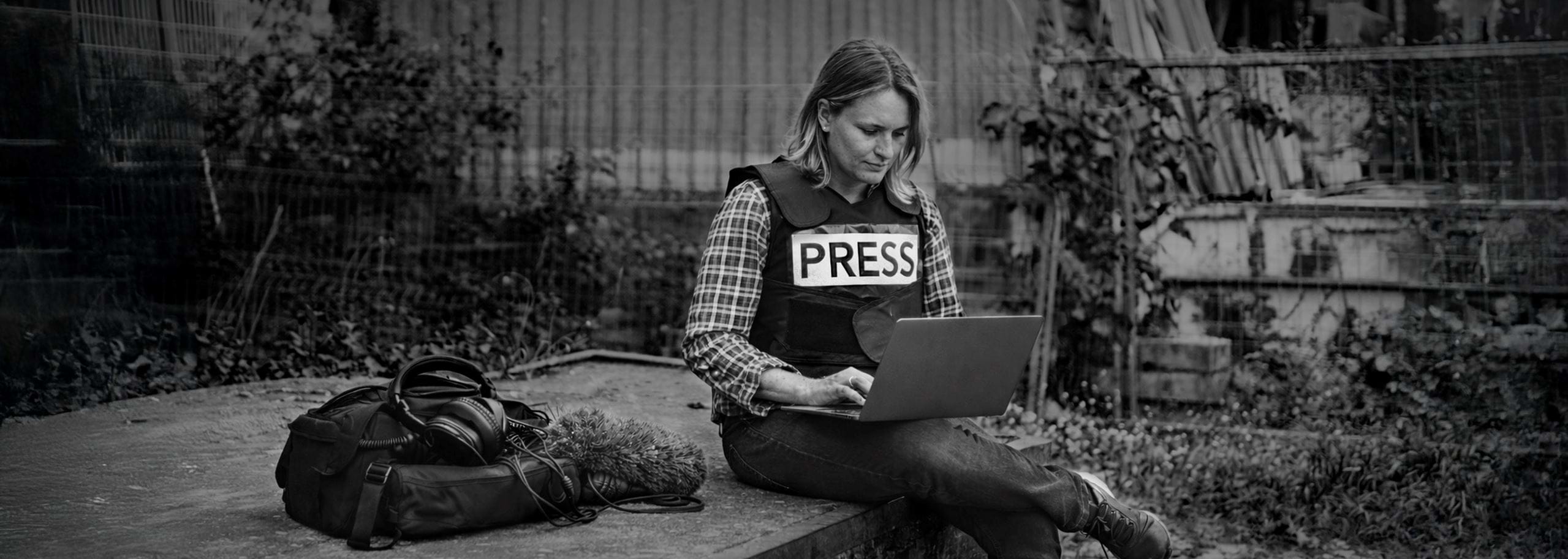 Female journalist wearing a press vest editing audio on a laptop using Hindenburg Pro in an outdoor conflict environment.