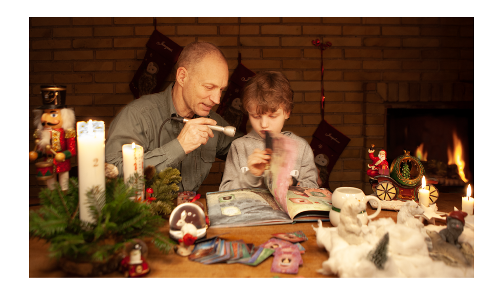 A journalist records a cozy Christmas scene at home, leaning in with a silver Electro Voice 635A microphone as a young boy flips through a picture book and explains his collection. The table is filled with holiday decorations, candles, small figurines, and scattered cards, while stockings hang on a brick wall and a fireplace glows in the background. The moment focuses on capturing both the child’s voice and the soft sounds of pages turning.