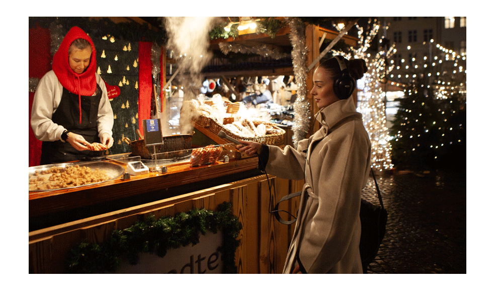 A female audio journalist records midrange ambience at a Christmas market food stall, wearing over-ear headphones and a beige winter coat. She extends a silver Electro-Voice 635A microphone toward the steaming grill as a vendor prepares food behind the counter, capturing sizzling, clattering utensils, and nearby crowd noise. Warm stall lights, garlands, and twinkling trees frame the scene, with wet cobblestones reflecting the festive glow.