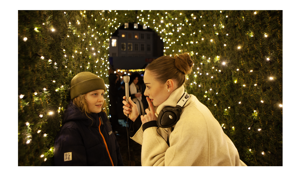 A female audio journalist leans toward a young boy inside a glowing tunnel of Christmas lights, holding a silver Electro-Voice 635A microphone between them. With a gentle finger to her lips, she reminds him—and the viewer—of the importance of capturing clean ambience for interviews, the subtle sound bed that fills gaps and smooths edits. Surrounded by warm festive lights and evergreen textures, the scene highlights quiet focus amid the bustle of the Christmas market.