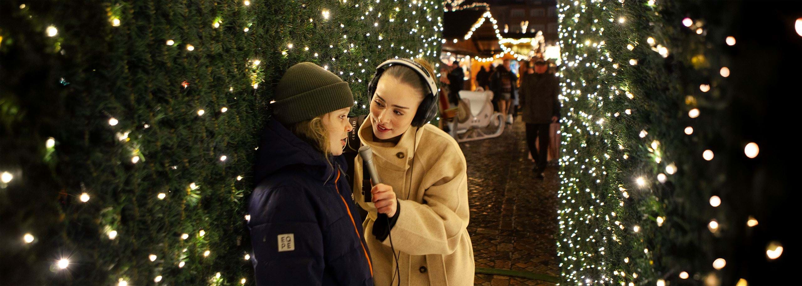 A female audio journalist leans in for an intimate, connected interview with a young boy, wearing over-ear headphones and holding a silver Electro-Voice 635A microphone close to capture his voice clearly. They’ve stepped away from the busiest parts of the Christmas market into a quieter passage framed by evergreen hedges wrapped in twinkling lights, creating a calm, private pocket of space. The noisy market fades into a soft, distant blur behind them.