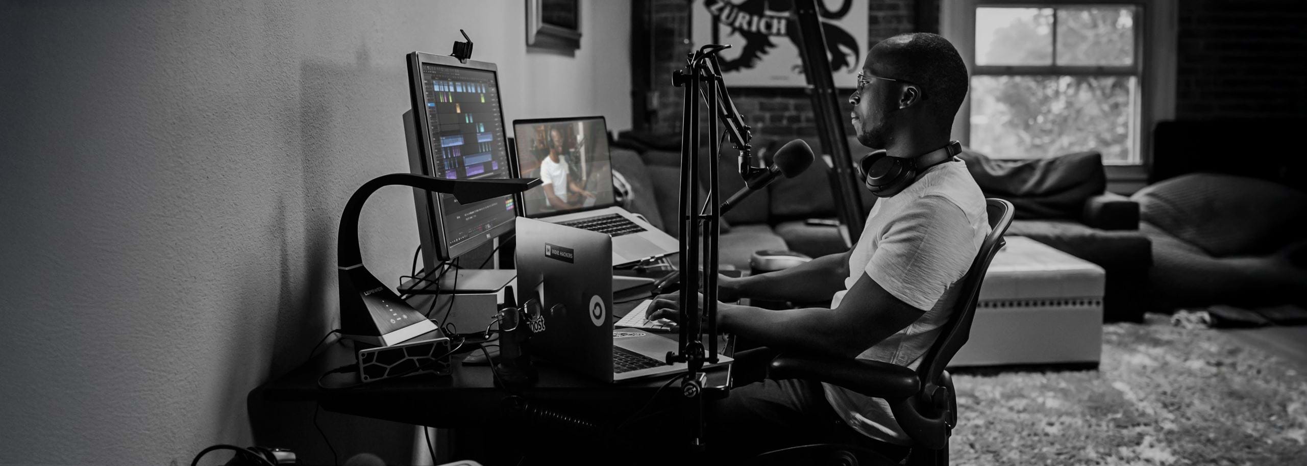 Journalist editing audio for video on a computer using Hindenburg Pro in a home studio setup with microphone and headphones.