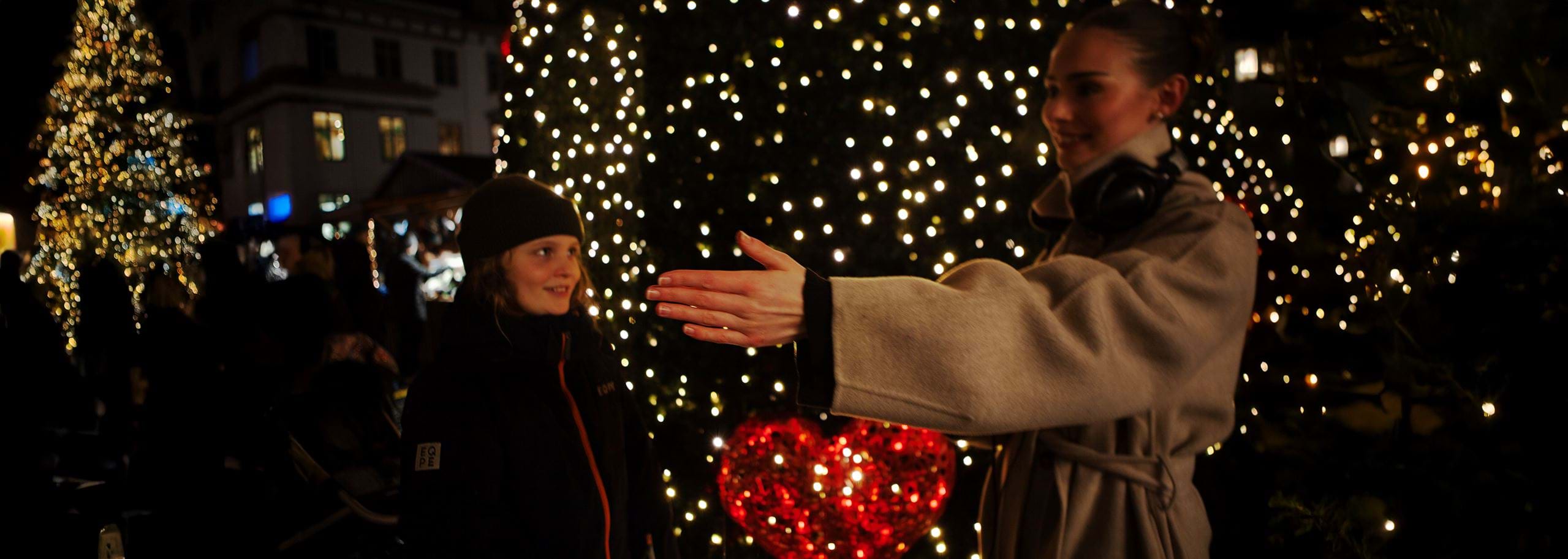 A female audio journalist at a Christmas market directs an interview, extending her arm to guide where the young boy should stand or speak. She wears a beige winter coat with headphones resting around her neck, focused and in control of the recording setup. The boy, in a dark jacket and beanie, looks toward her as they stand in front of a hedge covered in twinkling lights, with a glowing red heart decoration and festive trees sparkling in the background.