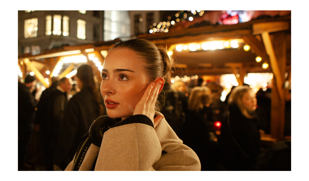 A female audio journalist stands in a crowded Christmas market under warm string lights and glowing wooden stalls. Wearing a beige winter coat with headphones resting around her neck, she presses one hand to a single ear—monitoring in mono to focus on balance and detail in the soundscape. Her gaze is fixed slightly upward and to the side, attentive and analytical, while the bustling crowd behind her blurs into soft golden bokeh, emphasizing the lively holiday atmosphere.