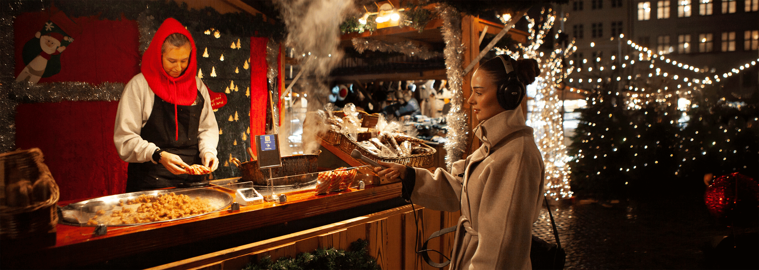 A female audio journalist records midrange ambience at a Christmas market food stall, wearing over-ear headphones and a beige winter coat. She extends a silver Electro-Voice 635A microphone toward the counter as a vendor prepares food beside a steaming grill, capturing sizzling sounds, utensil clatter, and nearby crowd noise. Festive garlands and warm lights frame the stall, while twinkling string lights and wet cobblestones glow in the background.