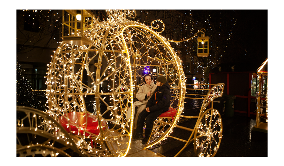 A wide nighttime view at a Christmas market shows a female audio journalist interviewing a young boy while seated inside a glowing, carriage-shaped holiday light installation. She wears over-ear headphones and a beige winter coat, holding a silver Electro-Voice 635A microphone toward him as they talk. The scene is filled with warm string lights, hanging lantern decorations, and twinkling trees, with wet pavement reflecting the festive glow in the background.