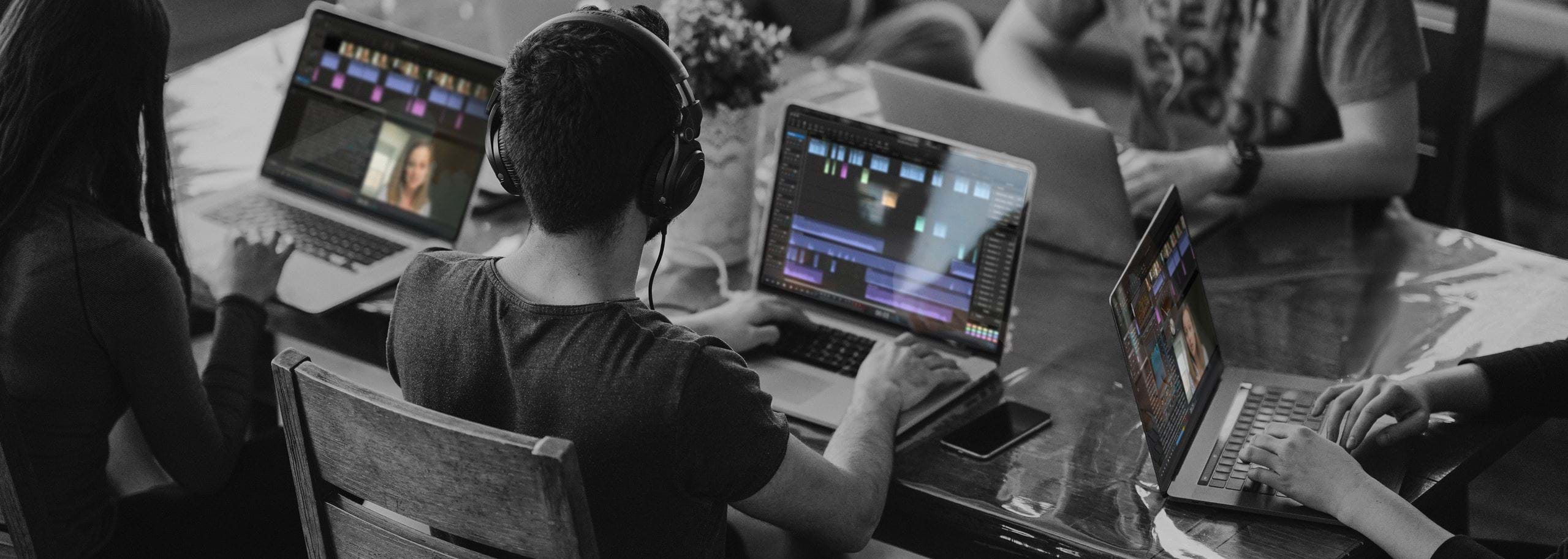 Students sitting around a table using laptops with audio editing timelines visible on their screens while one person listens with headphones.
